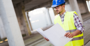 Engenheiro avalia projeto de estacionamento em obra de construção civil, usando capacete de segurança e vestimenta refletiva.
