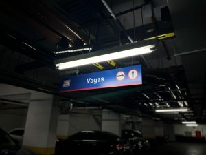 Placa de sinalização no estacionamento rotativo corporativo indicando vagas disponíveis, com setas para entrada e saída, iluminação de luzes fluorescentes e carros estacionados.