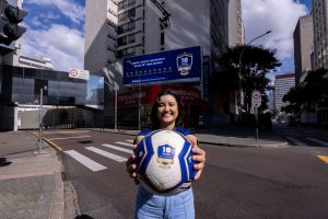 Foto de campanha em frente ao prédio da Invespark com cenário urbano ao fundo: uma mulher sorri segurando uma bola de futebol azul e branca com a marca “10 Copas de História Autopark”