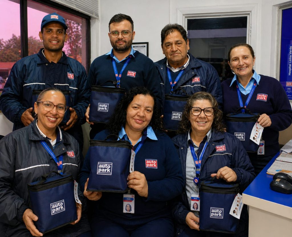 Equipe autopark reunida em evento, com profissionais uniformizados e credenciais, sorrindo na foto em frente a um balcão e porta de escritório.