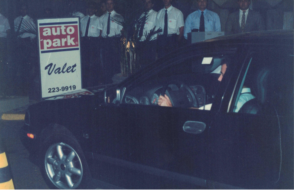 Registro das equipes no início dos anos 2000 da Valet Autopark, com funcionário uniformizado ao lado de placa “Autopark Valet” e carro em primeiro plano.