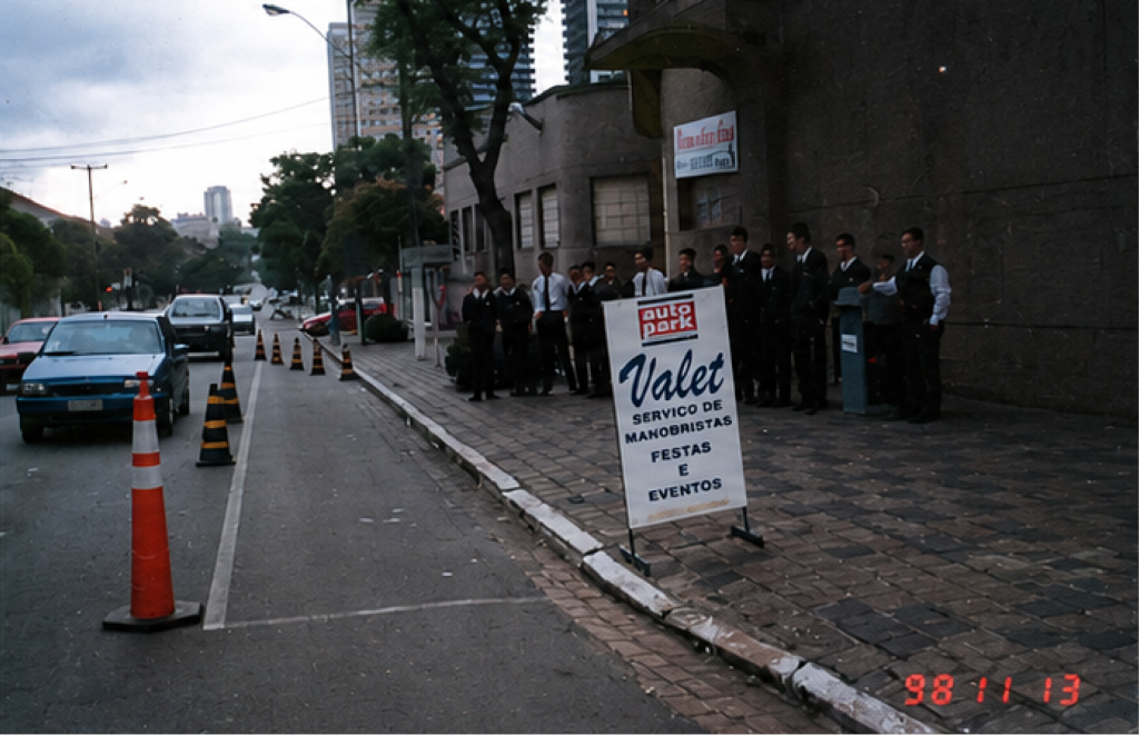 Registro das equipe em 1998 de Valet da Autopark, com profissionais em fila ao lado de veículos e cones laranja em via urbana, em frente a um prédio.