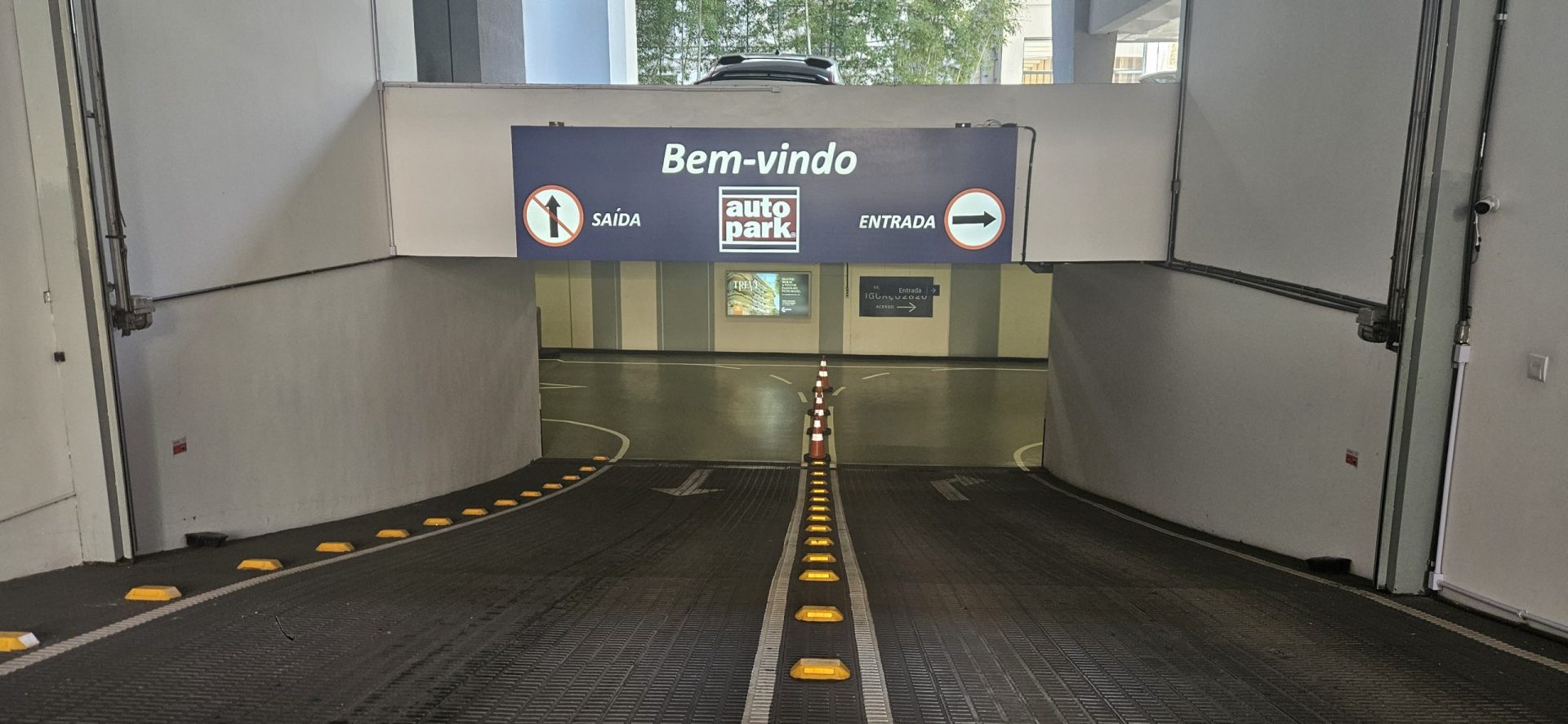 Entrada e saída sinalizadas em um estacionamento com faixa de orientação no chão e placas “Bem-vindo”, “Autopark”, “Entrada” e “Saída”, destacando gestão terceirizada de estacionamento e uma empresa de estacionamento.