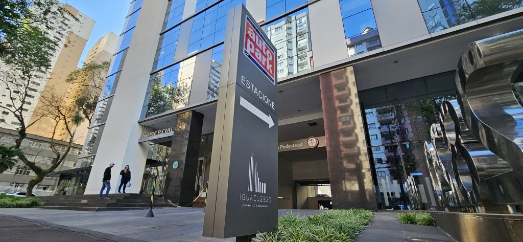 Prédio comercial em ambiente urbano com placa de estacionamento e acesso para veículos, mostrando indicações de tarifas e entrada no local