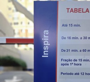 Placa de tabela de preço de estacionamento em estrutura azul e vermelha, com informações de tempo de permanência e valores para diferentes períodos.