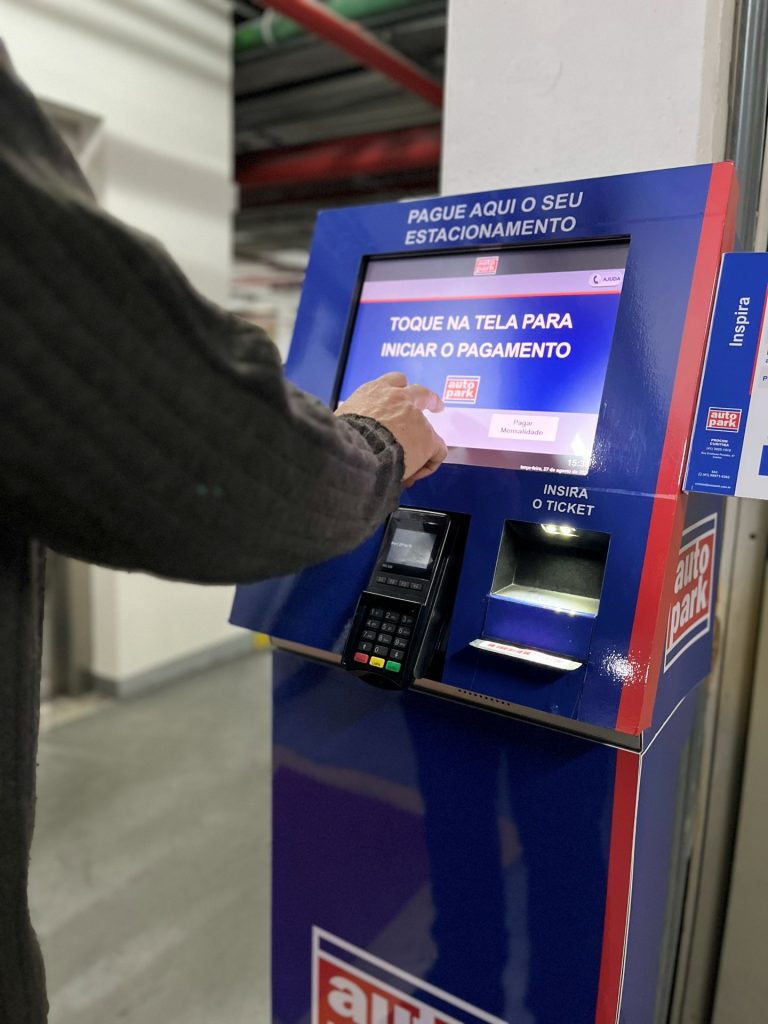 Pessoa usando um terminal de pagamento de estacionamento em autoatendimento, com tela orientando “Toque na tela para iniciar o pagamento”.