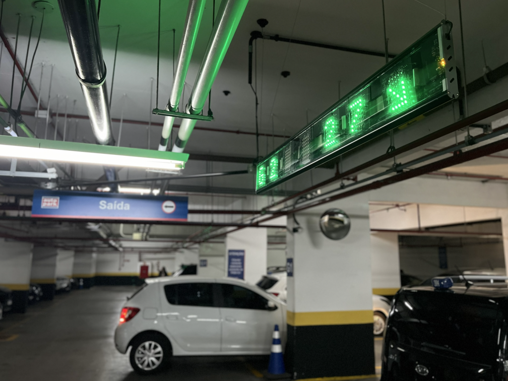 Estacionamento coberto com carro em vaga, sinalização de saída ao fundo e luzes verdes de controle no teto, em ambiente industrial de estacionamento.