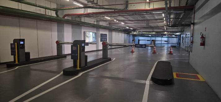 Corredor interno de estacionamento com baias demarcadas no piso, equipamentos de controle de acesso e cones de sinalização ao fundo, com placas de direção e áreas de circulação em ambiente industrial. 