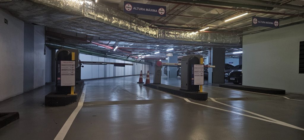Visão interna de um estacionamento rotativo em shopping, com vagas demarcadas no piso, barreiras de controle e placas de orientação em um corredor coberto.