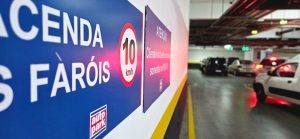 Corredor de estacionamento em shopping com veículos ao fundo e faixa de sinalização de velocidade e pedestres ao lado, indicando controle e organização do fluxo no local.
