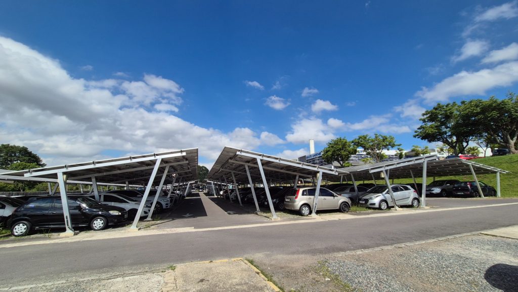 Estacionamento em shopping com vagas cobertas por estrutura metálica e diversos veículos ao fundo sob céu azul, mostrando a gestao de estacionamento em shopping em operação.