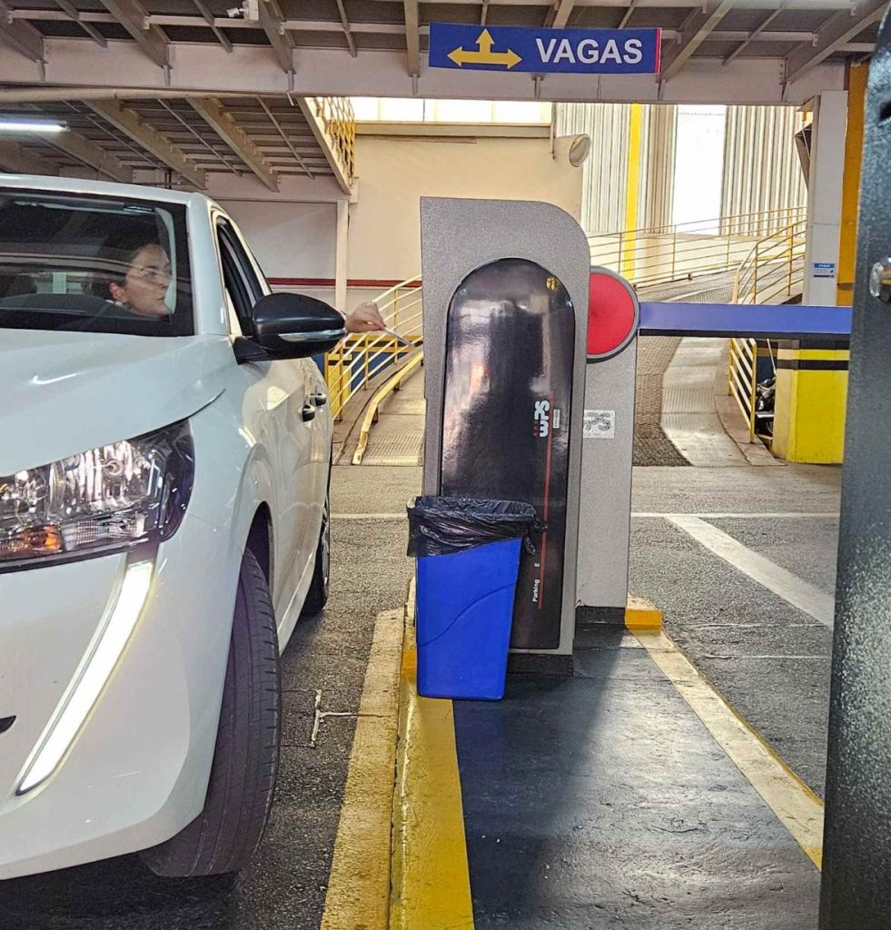 Vista interna de área de estacionamento em shopping com carro branco estacionado, faixas amarelas no piso e placa de sinalização de vagas com seta e identificação VAGAS.