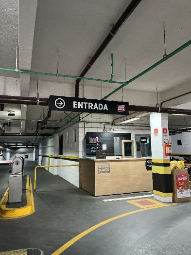 Plataforma de entrada de estacionamento em shopping, com placa “ENTRADA”, cabine de atendimento e sinalização de caixas, indicando fluxo automatizado de vagas.