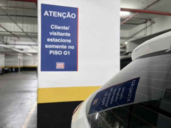 Sinal de atenção em estacionamento de shopping com orientações para clientes, destacando que visitantes estacionem apenas no piso G1. Veículo com adesivo de autorização.