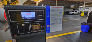 Painel e totem de atendimento em estacionamento com tela de controle e credenciamento, ao lado de tabela de preços, em área interna de shopping, com carros ao fundo
