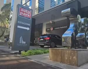 Placa indicativa de estacionamento rotativo comercial com seta e sinalização de segurança em Autopark, em frente a fachada de edifício comercial, com carros no acesso.