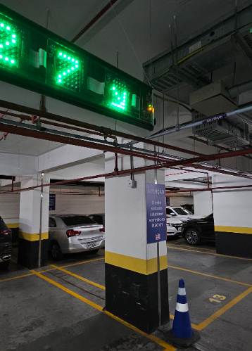 Estacionamento rotativo comercial em garagem: área com vagas demarcadas, cones de sinalização, carros parados e painel luminoso verde ao fundo.