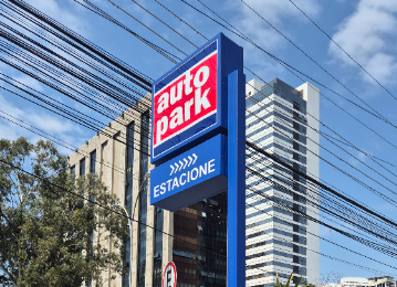 Placa de estacionamento rotativo comercial em área urbana com indicação “Estacionamento” ao lado de grades azuis e veículos em via comercial, com prédios ao fundo.