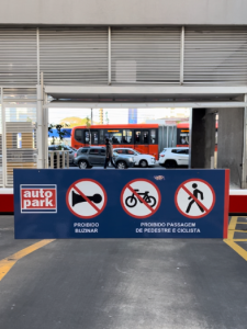 Placa de sinalização em área de entrada com advertências de trânsito, incluindo “Proibido buzinar” e “Proibida passagem de pedestre e ciclista” em estacionamento terceirizado Autopark, com carros e ônibus ao fundo.