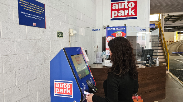 Pessoa atendendo em balcão de estacionamento terceirizado com placa “Autopark” em um prédio comercial, ao lado de caixas e sinalização de acesso.