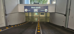 Entrada de estacionamento comercial terceirizado com a Autopark com sinalização “Bem-vindo”, faixas de direção e vagas demarcadas, vista do corredor central em garagem coberta.