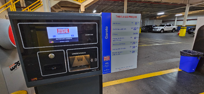 Painel de controle em estacionamento comercial terceirizado com tela de informações, opção de credenciamento e tabela de preços ao fundo, em área de pátio com vagas demarcadas.