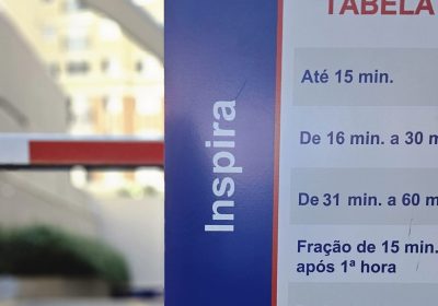 Placa de tabela de preço de estacionamento em estrutura azul e vermelha, com informações de tempo de permanência e valores para diferentes períodos.