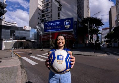 Foto de campanha em frente ao prédio da Invespark com cenário urbano ao fundo: uma mulher sorri segurando uma bola de futebol azul e branca com a marca “10 Copas de História Autopark”
