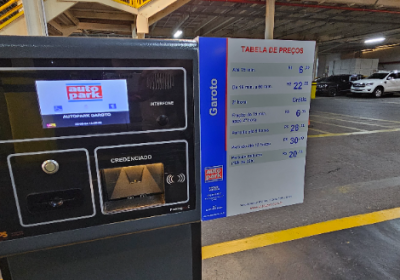 Painel e totem de atendimento em estacionamento com tela de controle e credenciamento, ao lado de tabela de preços, em área interna de shopping, com carros ao fundo