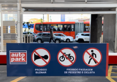 Placa de sinalização em área de entrada com advertências de trânsito, incluindo “Proibido buzinar” e “Proibida passagem de pedestre e ciclista” em estacionamento terceirizado Autopark, com carros e ônibus ao fundo.