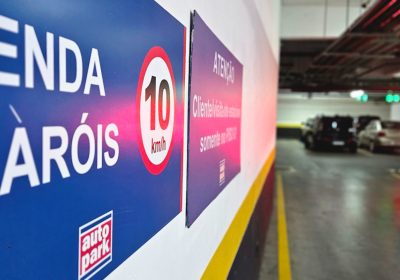 Corredor de estacionamento em shopping com veículos ao fundo e faixa de sinalização de velocidade e pedestres ao lado, indicando controle e organização do fluxo no local.