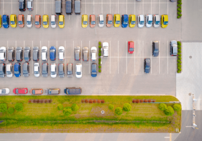 Imagem aérea mostrando amplo estacionamento de shopping com várias vagas disponíveis, áreas verdes e entradas de veículos,