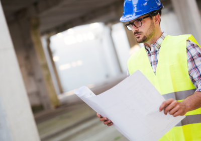Engenheiro avalia projeto de estacionamento em obra de construção civil, usando capacete de segurança e vestimenta refletiva.