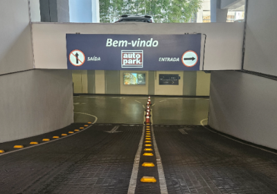 Entrada de estacionamento comercial terceirizado com a Autopark com sinalização “Bem-vindo”, faixas de direção e vagas demarcadas, vista do corredor central em garagem coberta.